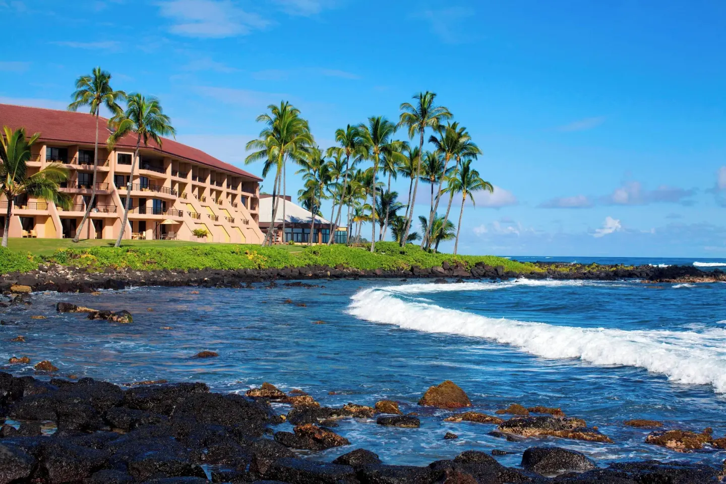Ocean waves and rocky shoreline beside Sheraton Kauai Resort with palms and guest buildings.