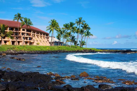 Ocean waves and rocky shoreline beside Sheraton Kauai Resort with palms and guest buildings.