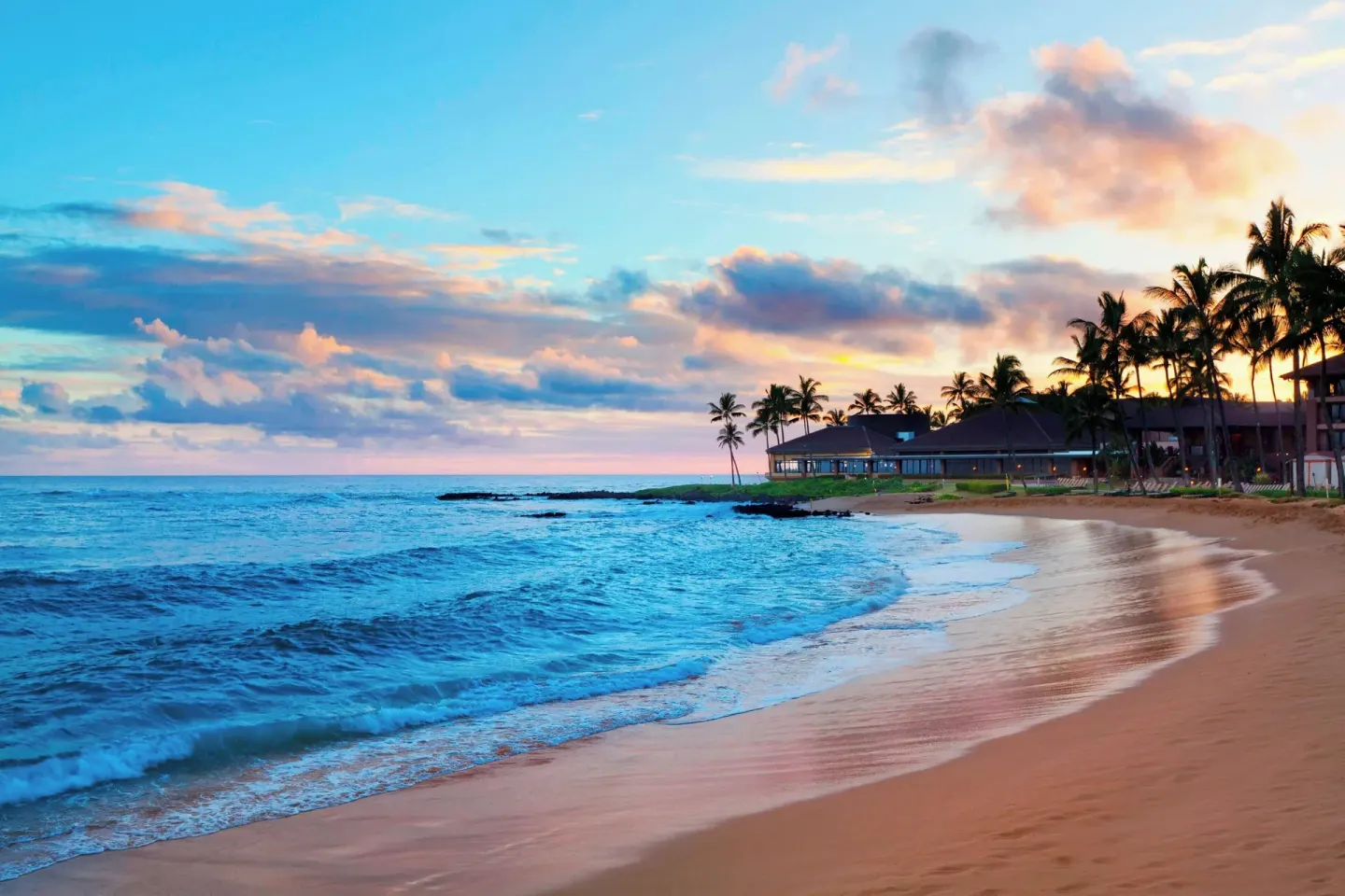 Sunset colors over the beach and ocean at Sheraton Kauai Resort with palms and resort buildings.