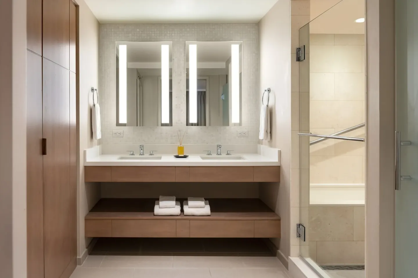 Bathroom with double sinks, lit mirrors, wood storage, and glass shower at Sheraton Waikiki Beach Resort.