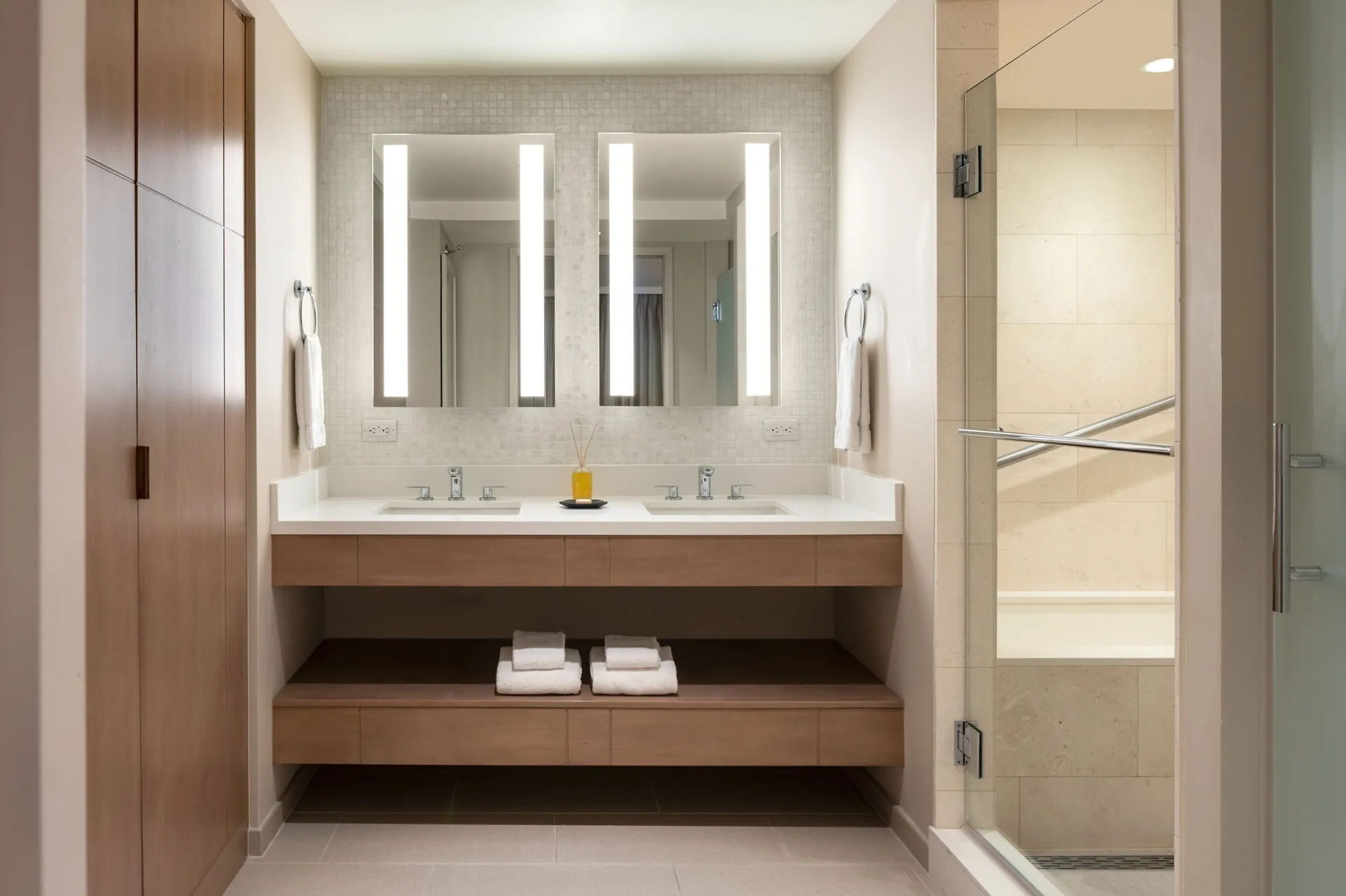 Bathroom with double sinks, lit mirrors, wood storage, and glass shower at Sheraton Waikiki Beach Resort.