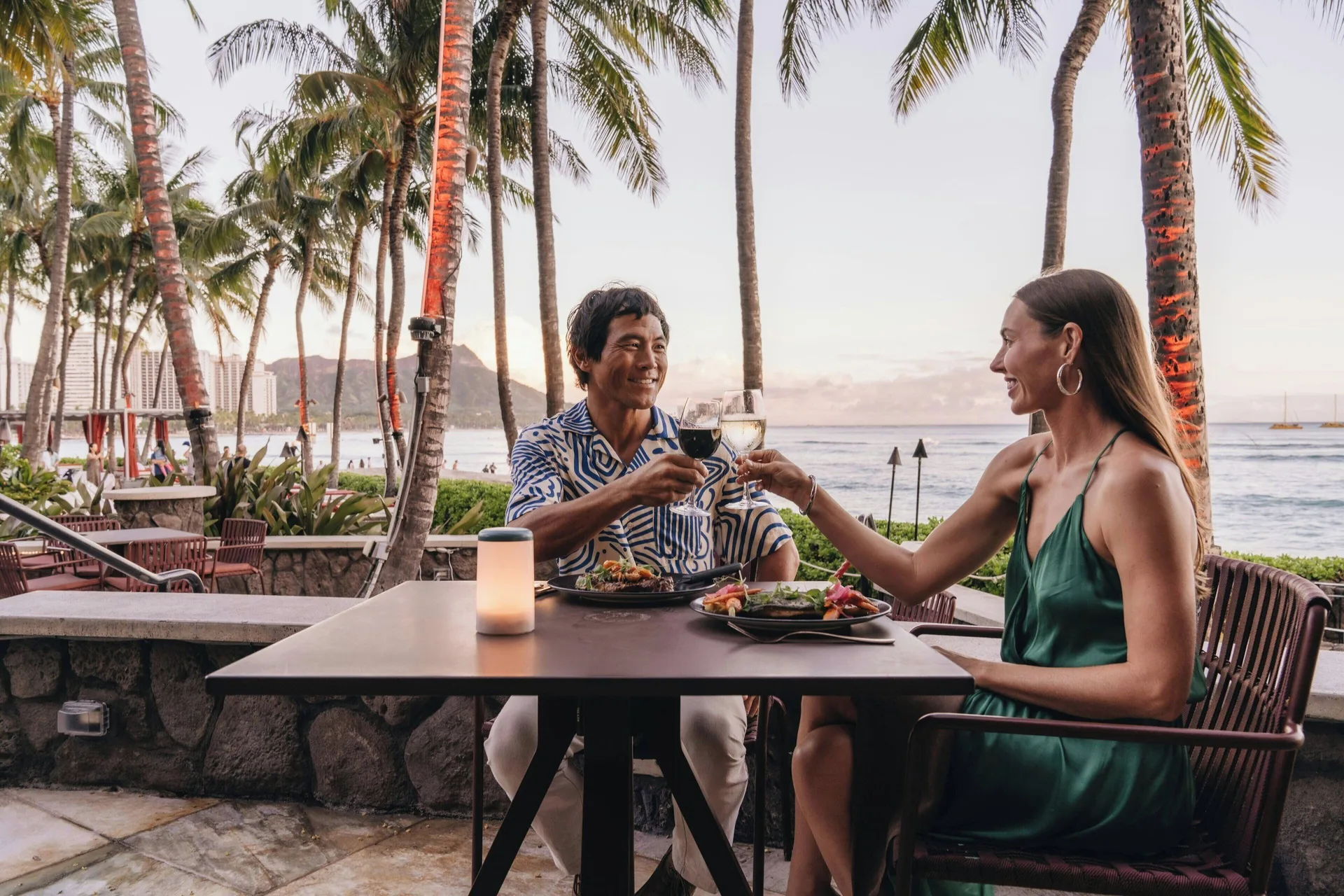 Oceanfront outdoor dining with two guests toasting at Sheraton Waikiki Beach Resort.
