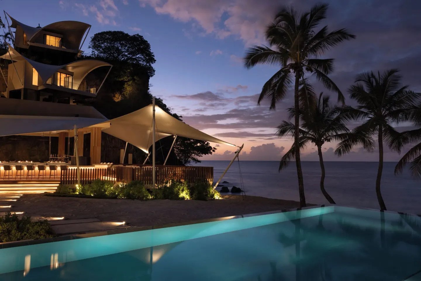 The illuminated pool, bar area, palms, and sea at dusk at Silversands Beach House Grenada.