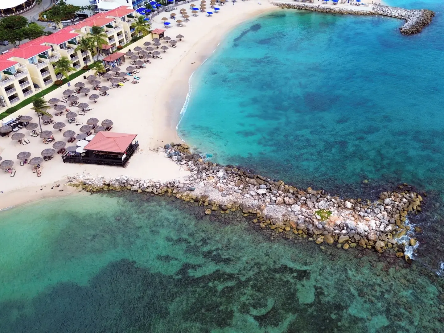 Aerial beach cove at Simpson Bay Resort, Marina & Spa with resort buildings, palapas, and clear water.