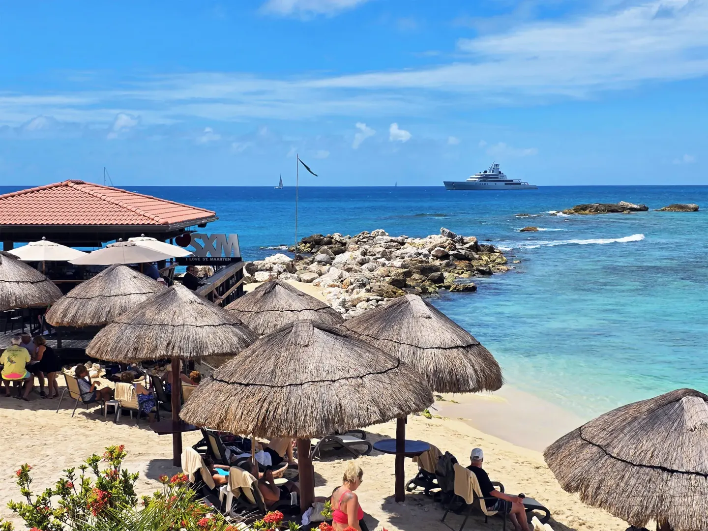 Beach palapas and ocean at Simpson Bay Resort, Marina & Spa with guests seated near a seaside bar area.