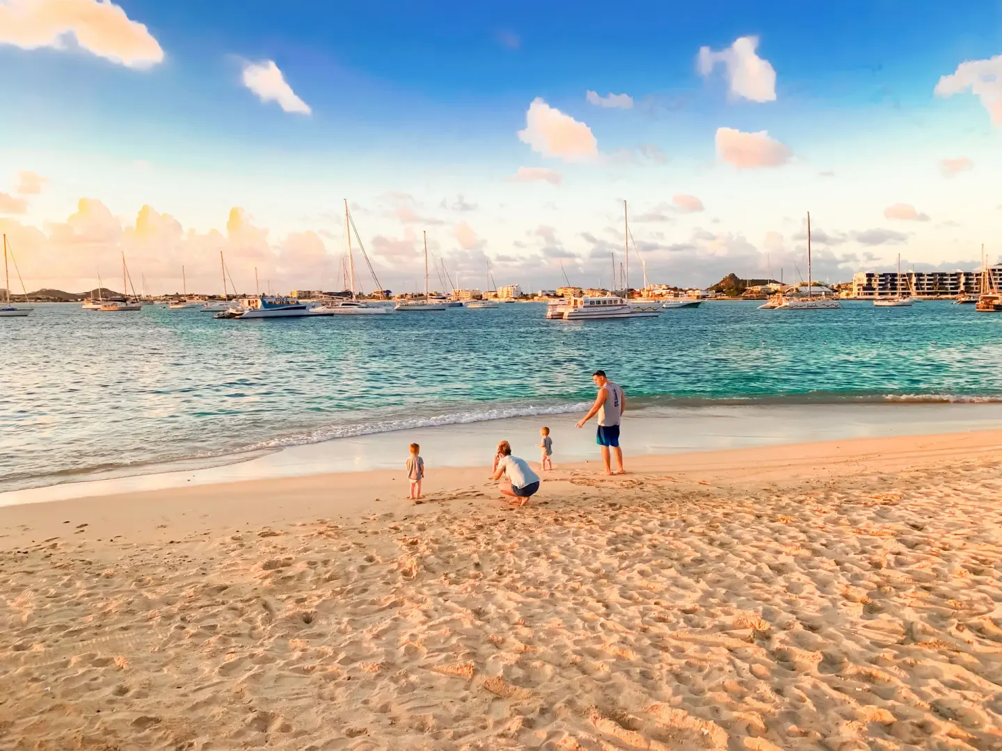 Family on the beach at Simpson Bay Resort, Marina & Spa with anchored boats across the bay at sunset.
