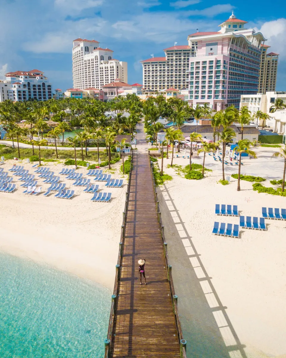 A beach pier leads from turquoise water toward SLS Baha Mar and palm trees.