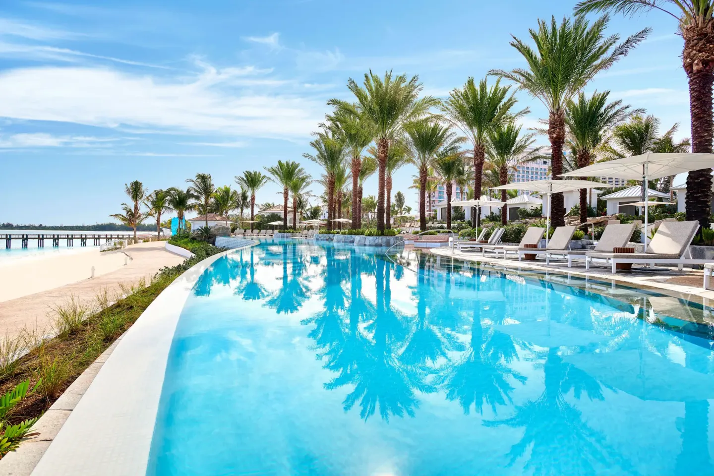 A palm-lined pool runs beside the beach with loungers and umbrellas at SLS Baha Mar.
