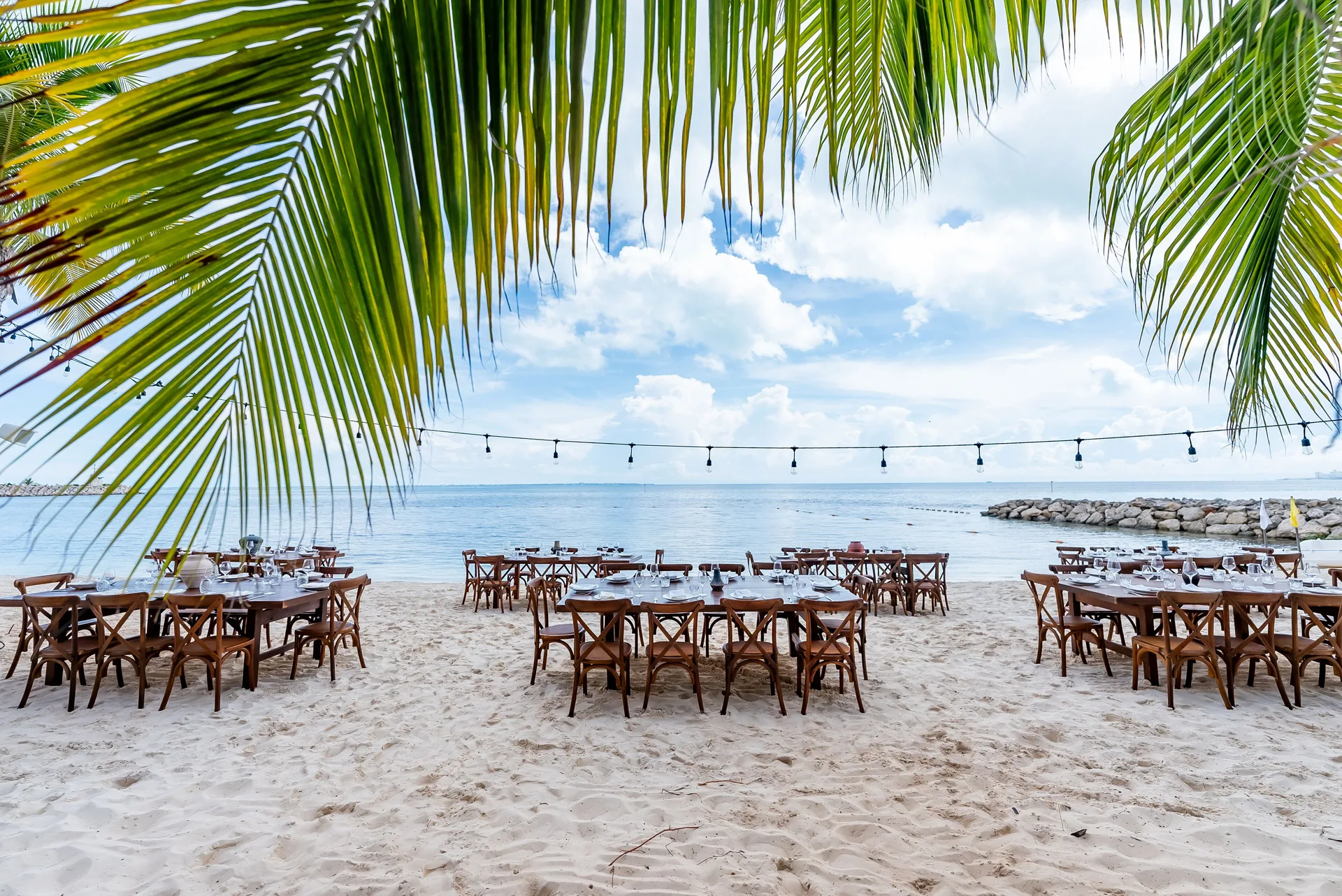 Beachfront dining tables are set on the sand beneath palm fronds at SLS Cancun.