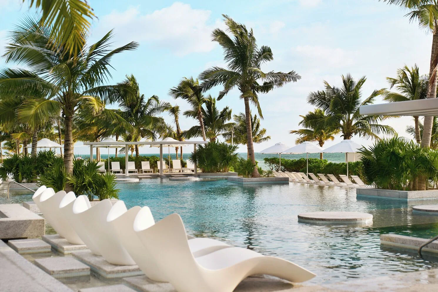 Resort pool with white loungers, palms, and ocean beyond at SLS Playa Mujeres.