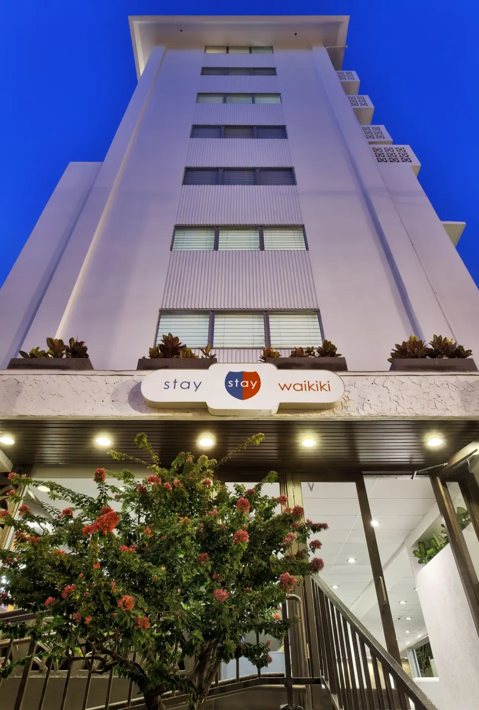 The Stay Hotel Waikiki exterior rises above its entrance sign at dusk.