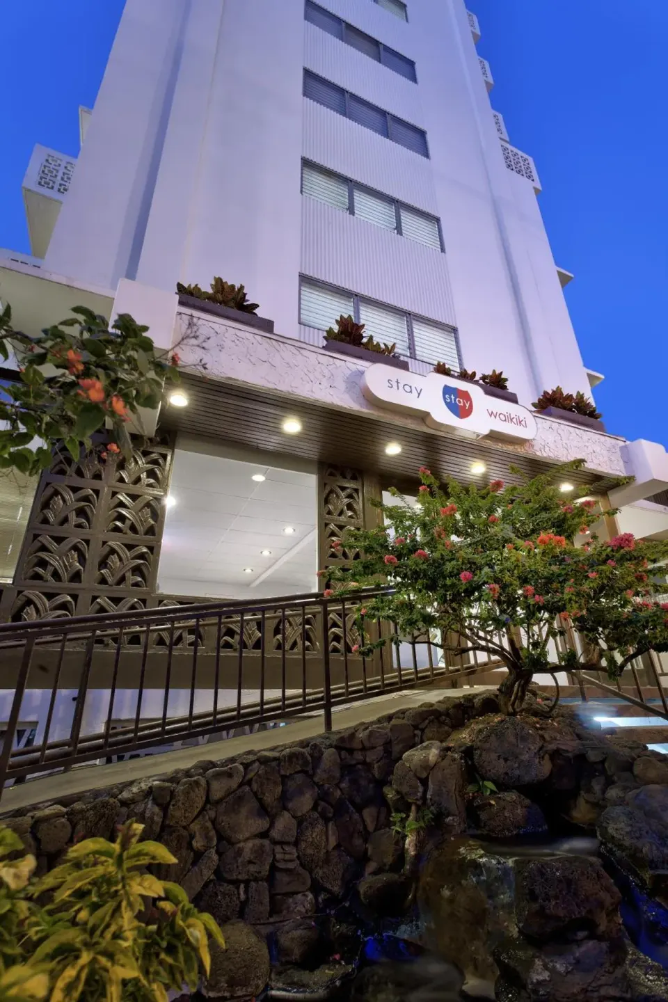 Stay Hotel Waikiki exterior with entrance sign, flowering plants, and rock waterfall at dusk.