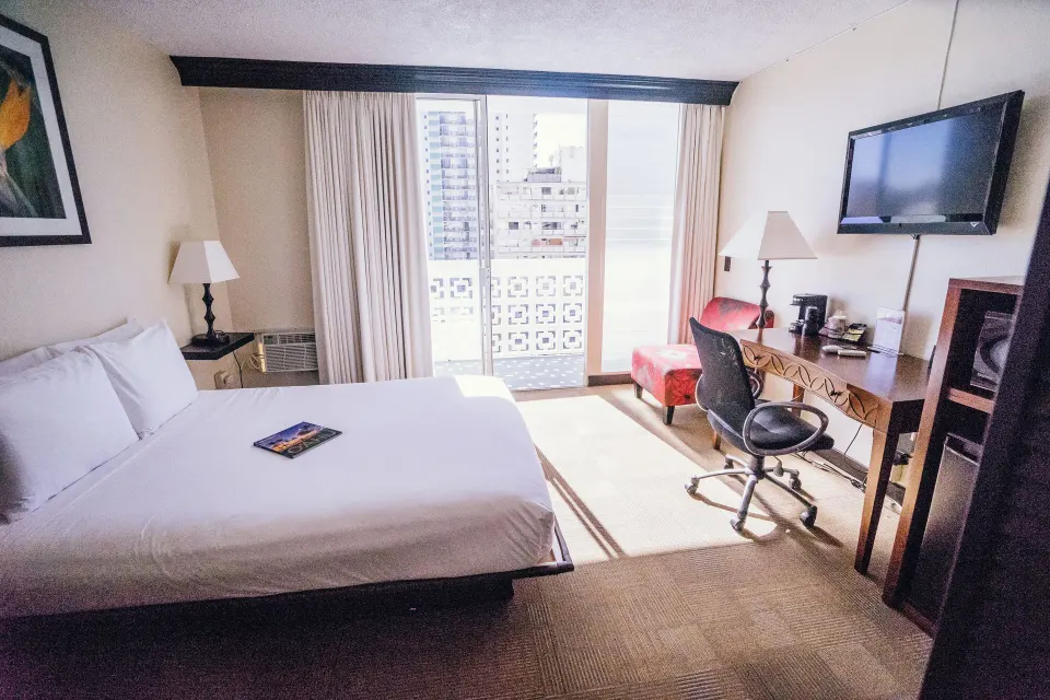 Petite Room with double bed, desk, wall TV, red chair, and balcony light at Stay Hotel Waikiki.