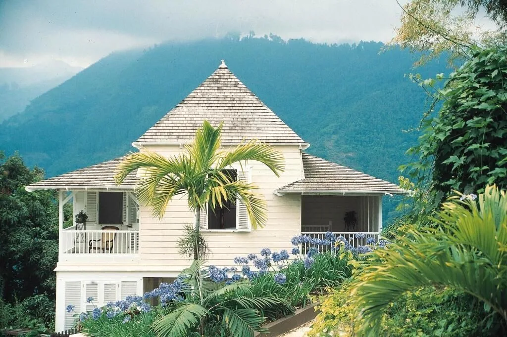 A white cottage at Strawberry Hill is framed by palms, flowers, and misty blue mountains.
