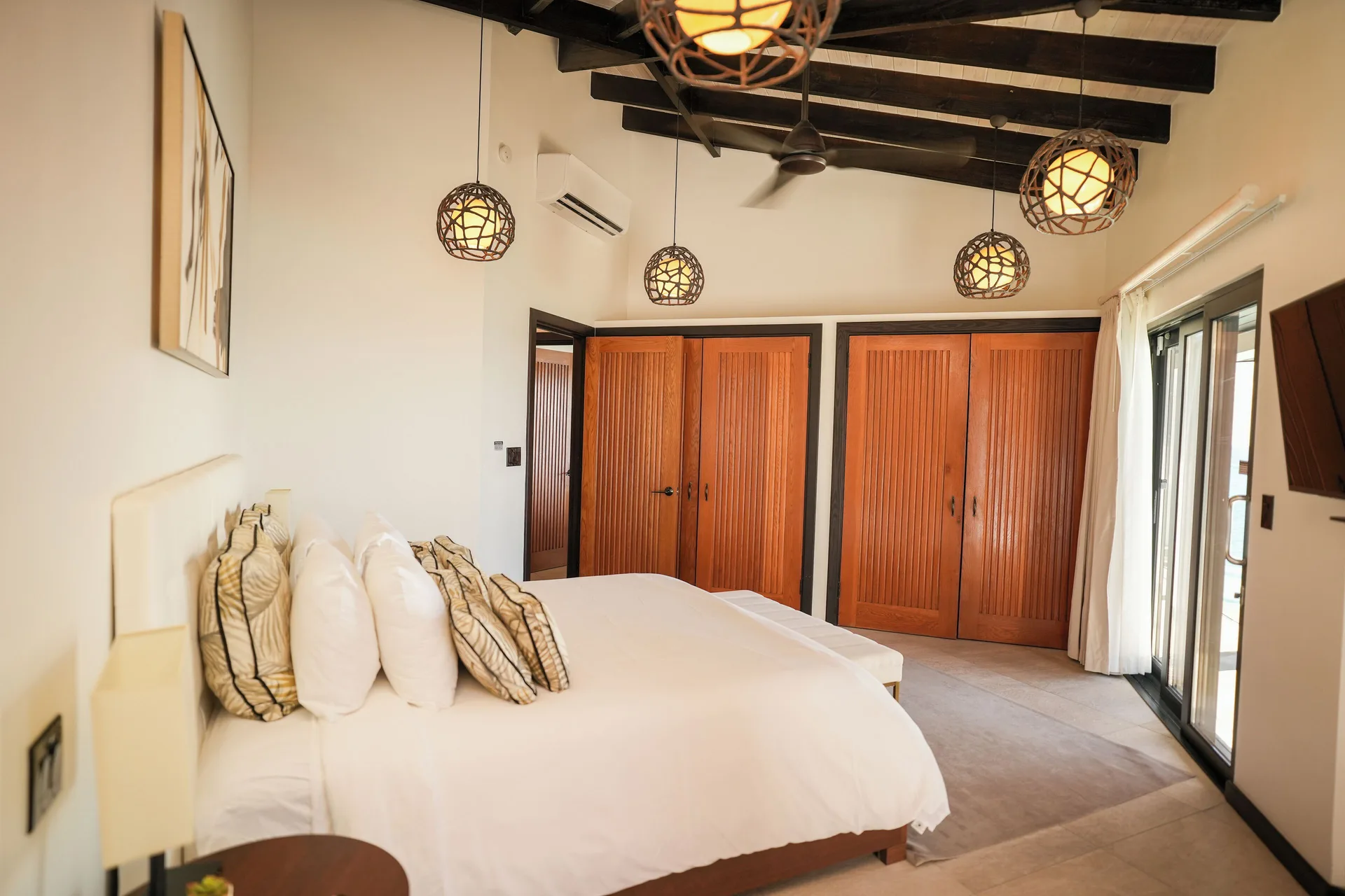 Bedroom with white bed, wood closets, pendant lights, and balcony doors at Tamarind Hills Resort & Villas.