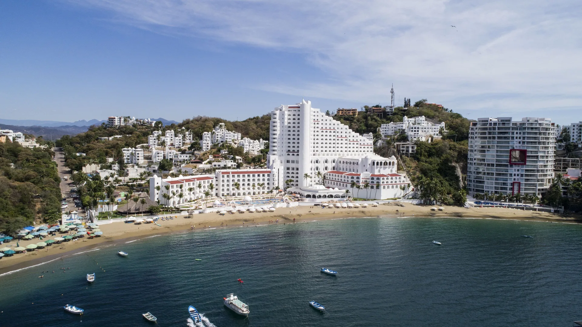 Wide aerial view of Tesoro Manzanillo beside a curved beach and blue bay with small boats.