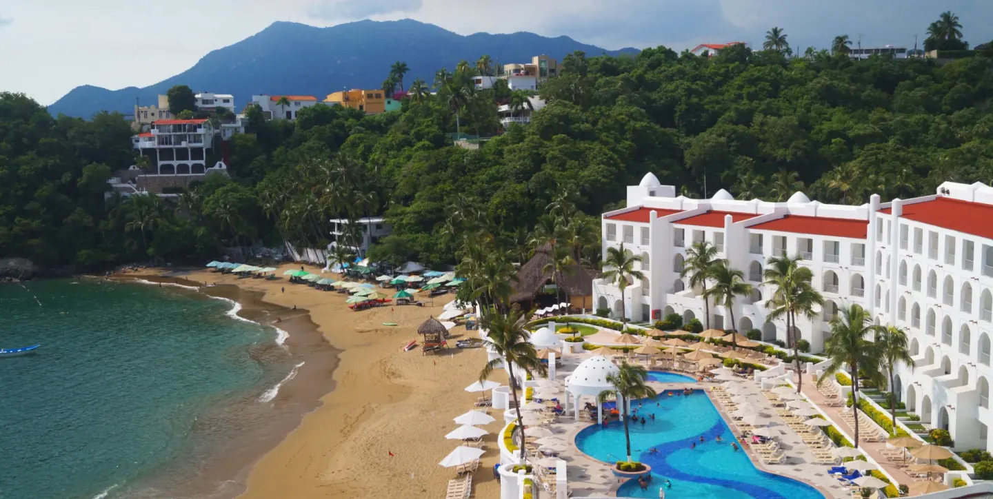 Beach and pool area at Tesoro Manzanillo with palm trees, loungers, and hillside greenery.
