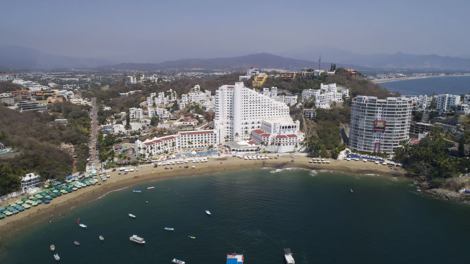 Aerial view of Tesoro Manzanillo on the beachfront with bay water, boats, and surrounding hills.