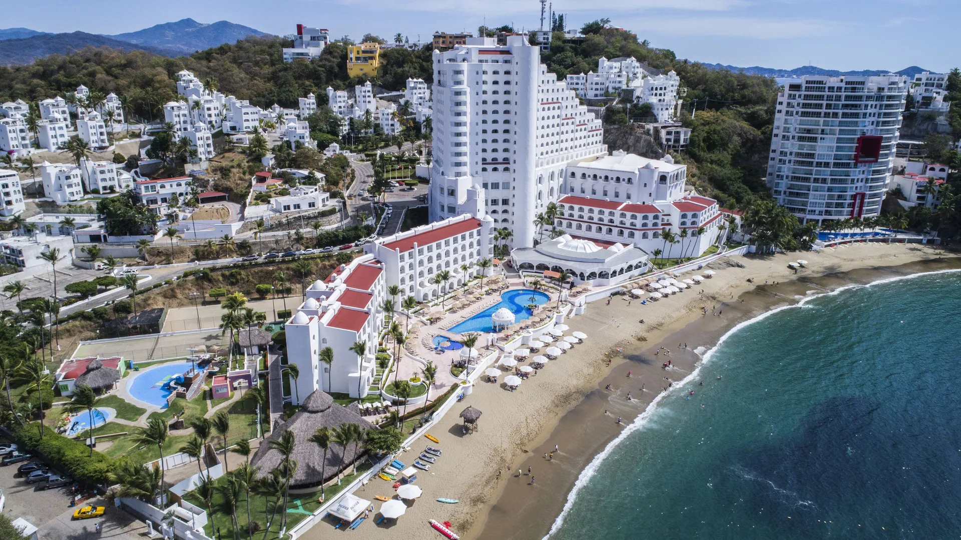 Angled aerial view of Tesoro Manzanillo with beachfront pool, beach umbrellas, and ocean shoreline.