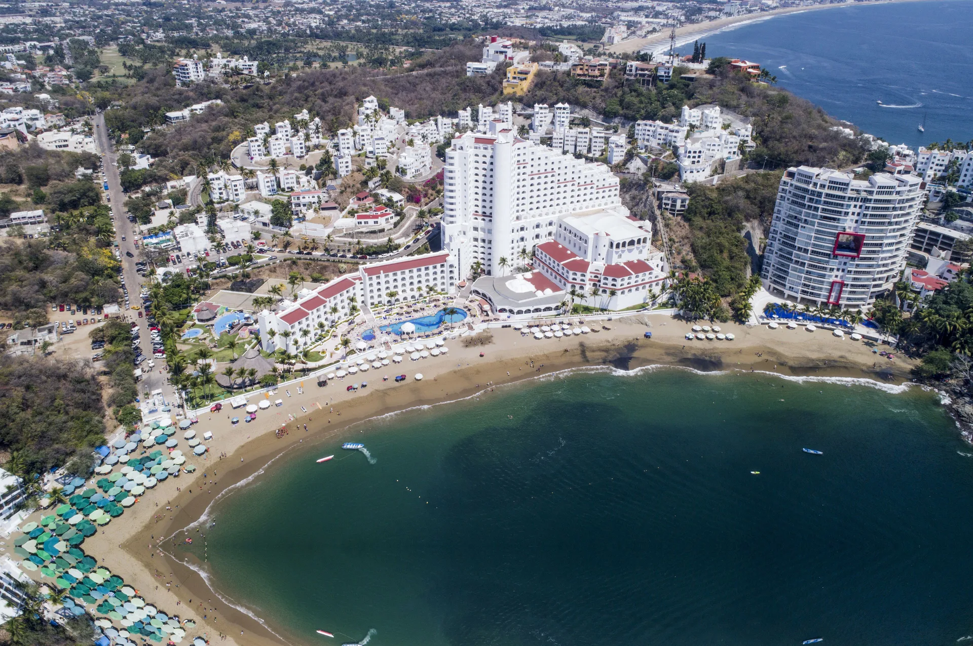 Aerial view of Tesoro Manzanillo, beach umbrellas, pools, and bay shoreline.