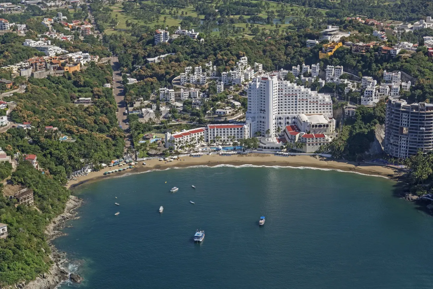 Wide aerial view of Tesoro Manzanillo on a cove with beach, boats, and hills.