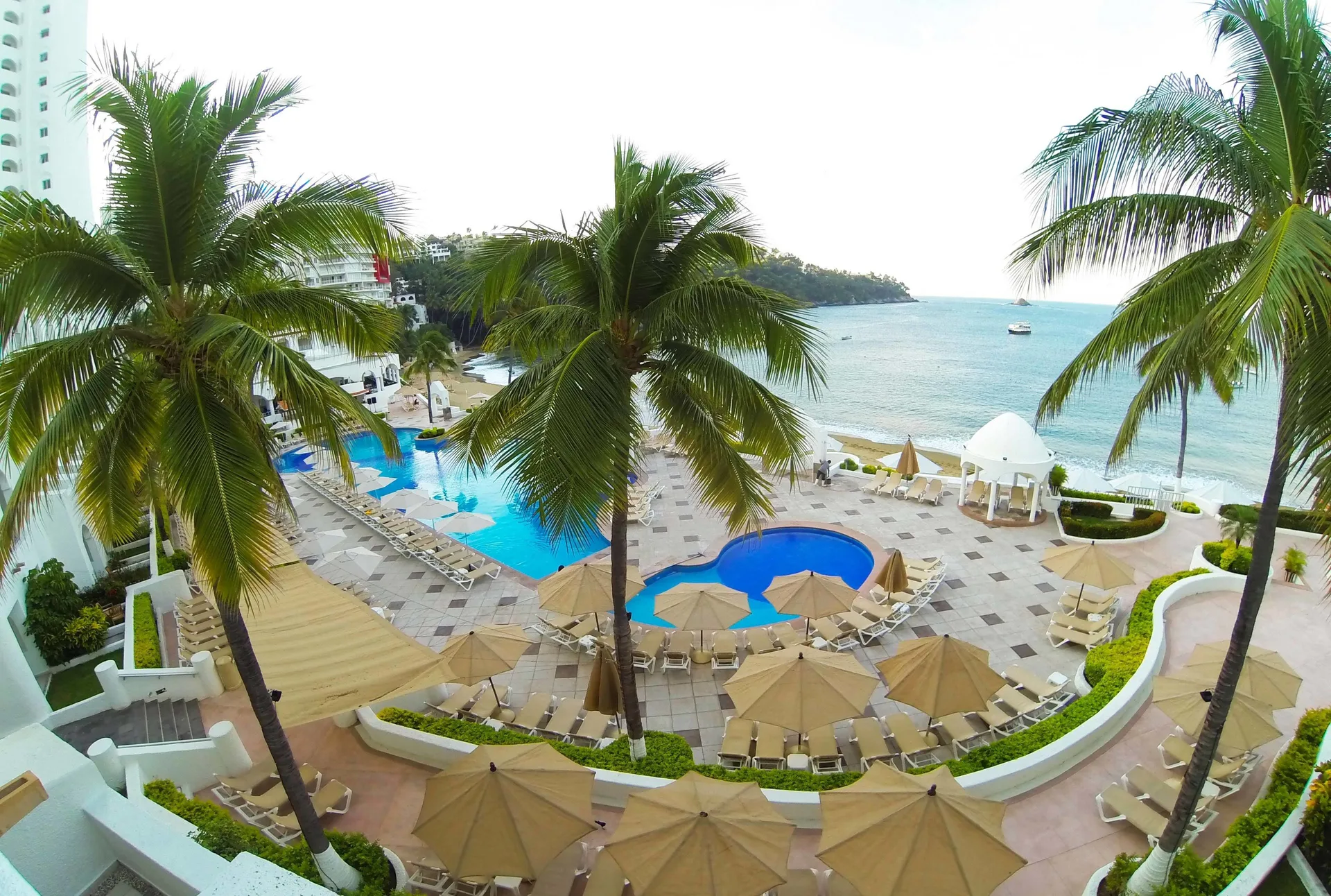 Pool terrace at Tesoro Manzanillo with palm trees, loungers, umbrellas, and ocean beyond.