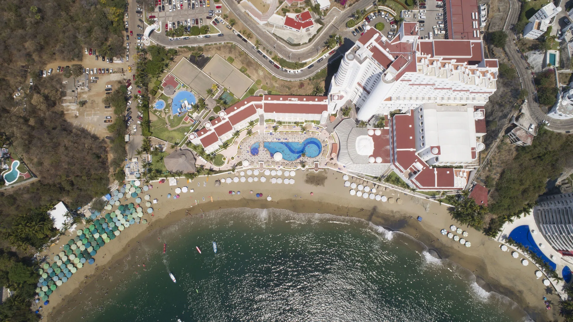 Top-down aerial of Tesoro Manzanillo with pools, beach, umbrellas, and waves.