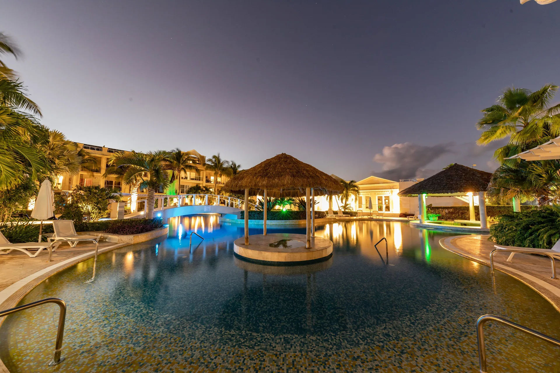 Evening pool area with palapas, loungers and illuminated water at The Atrium Resort.