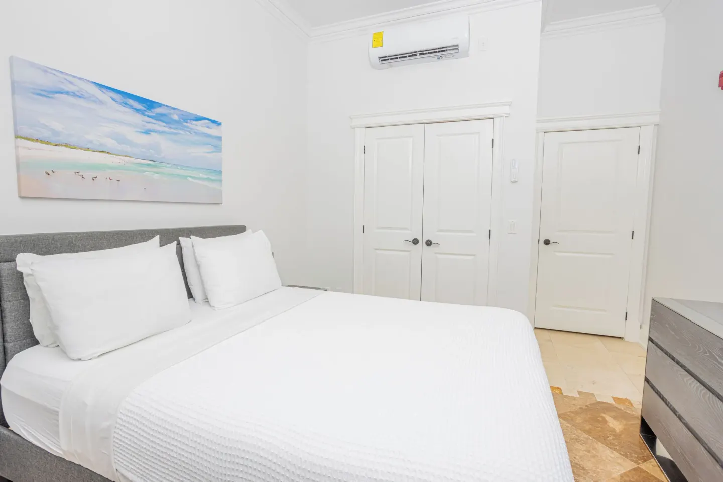 Guest room bed with white linens, beach artwork, closet doors, and dresser at The Atrium Resort.