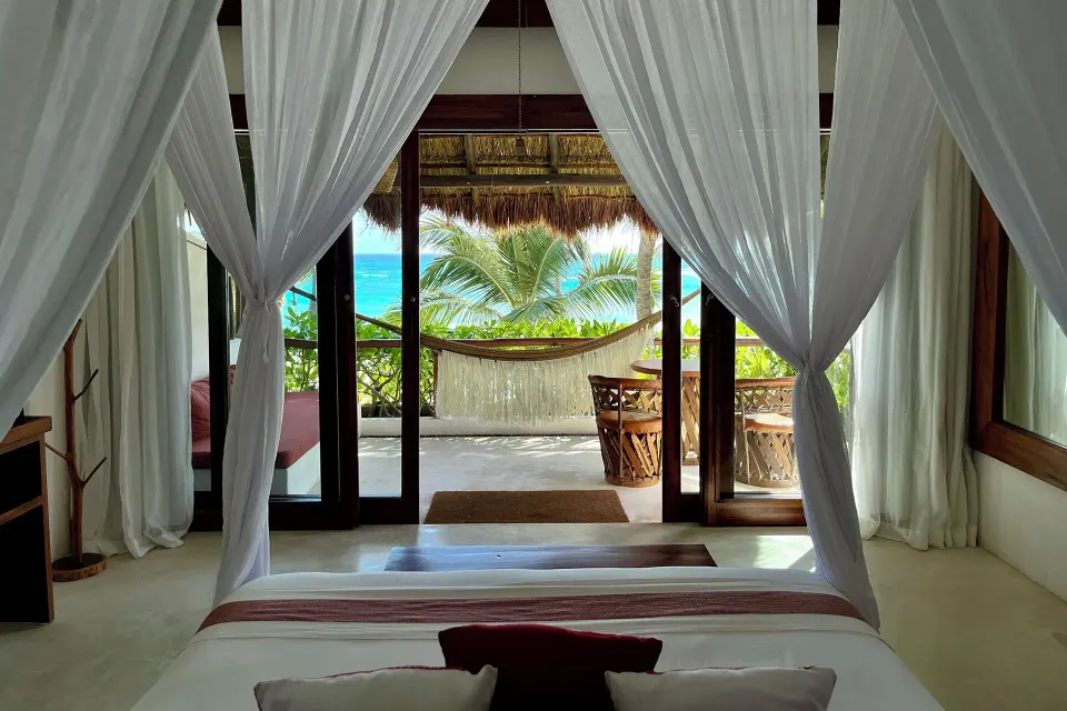 View from a canopy bed through glass doors to a hammock and ocean at The Beach Tulum