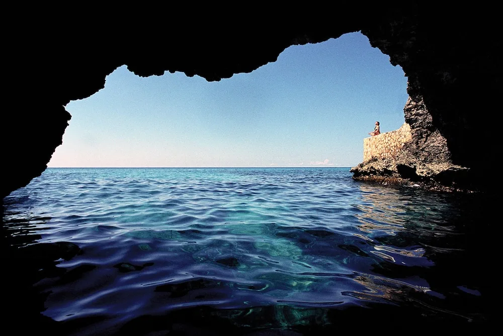 Sea cave at The Caves framing blue ocean, open sky, and a stone cliff platform with a seated person.