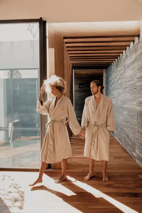Couple in robes holding hands in a sunlit spa hallway at The Club at Solaz.