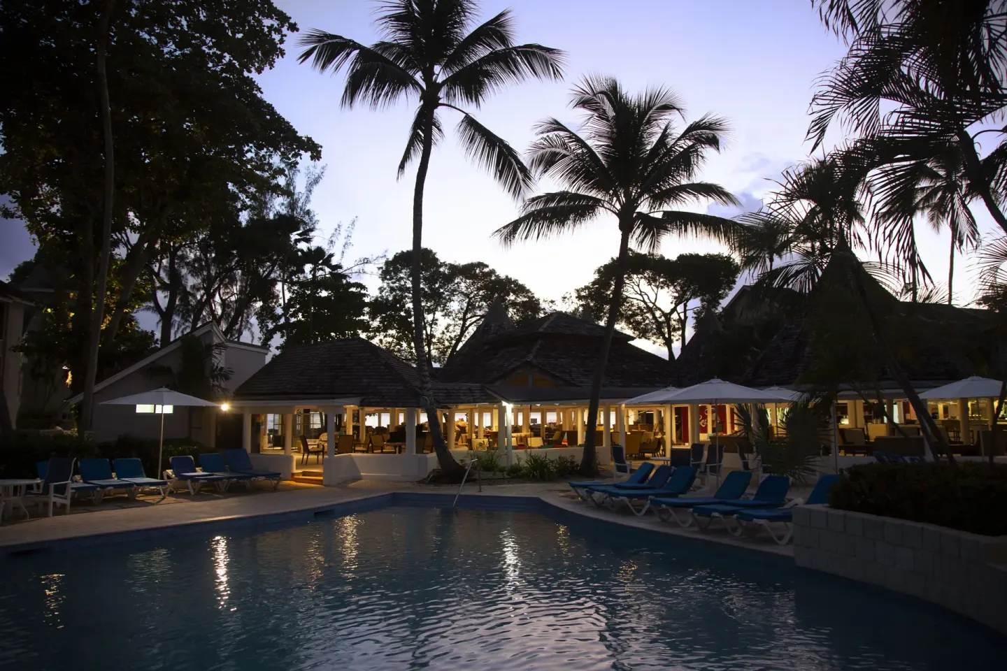 Pool with palm silhouettes and lit dining pavilion at twilight at The Club, Barbados Resort & Spa.