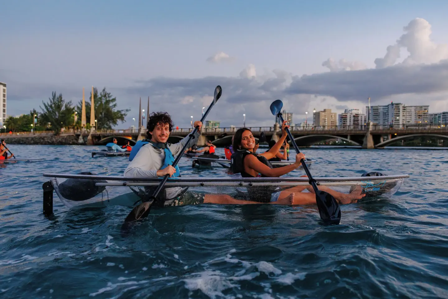 Two guests smile from a clear kayak near The Condado Plaza Hotel waterfront.