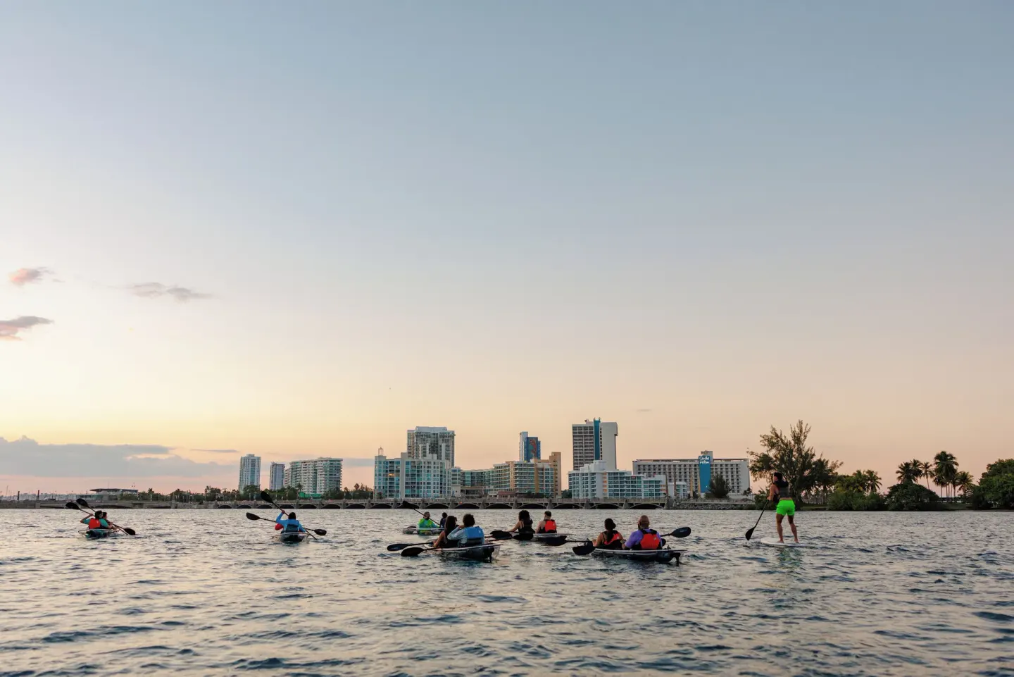 Kayakers and a paddleboarder gather on the lagoon near The Condado Plaza Hotel at dusk.