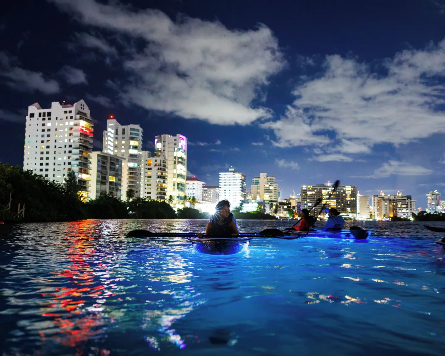 Night kayakers paddle on the lagoon near The Condado Plaza Hotel and city lights.
