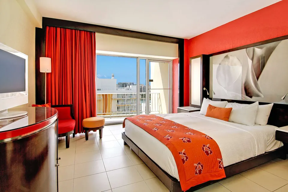 Deluxe Room with king bed, red accents, TV, chair, and balcony at The Condado Plaza Hotel.
