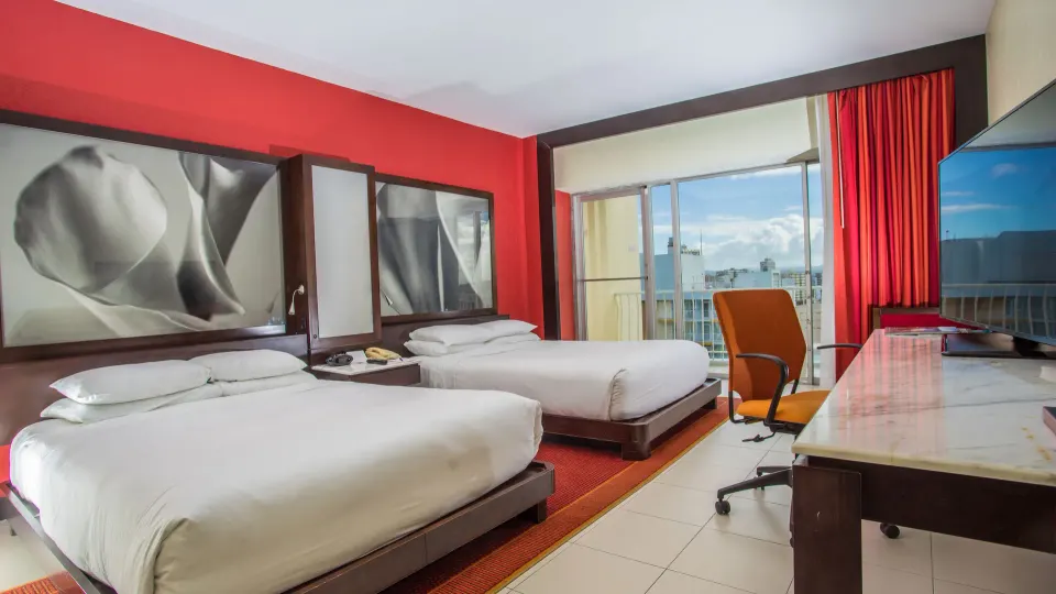 Deluxe Room with two beds, desk, TV, red wall, and balcony at The Condado Plaza Hotel.