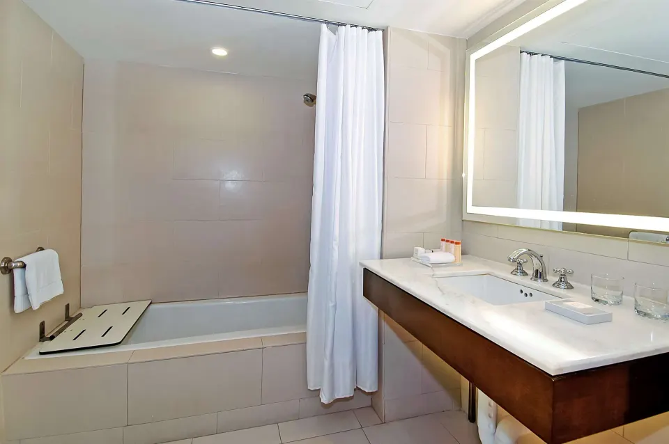 Accessible bathroom with bathtub, shower curtain, fold-down seat, and vanity at The Condado Plaza Hotel.