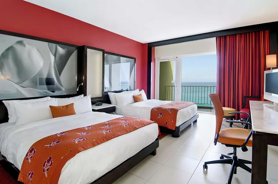 Guest room with two beds, red accents, desk, balcony, and ocean view at The Condado Plaza Hotel.