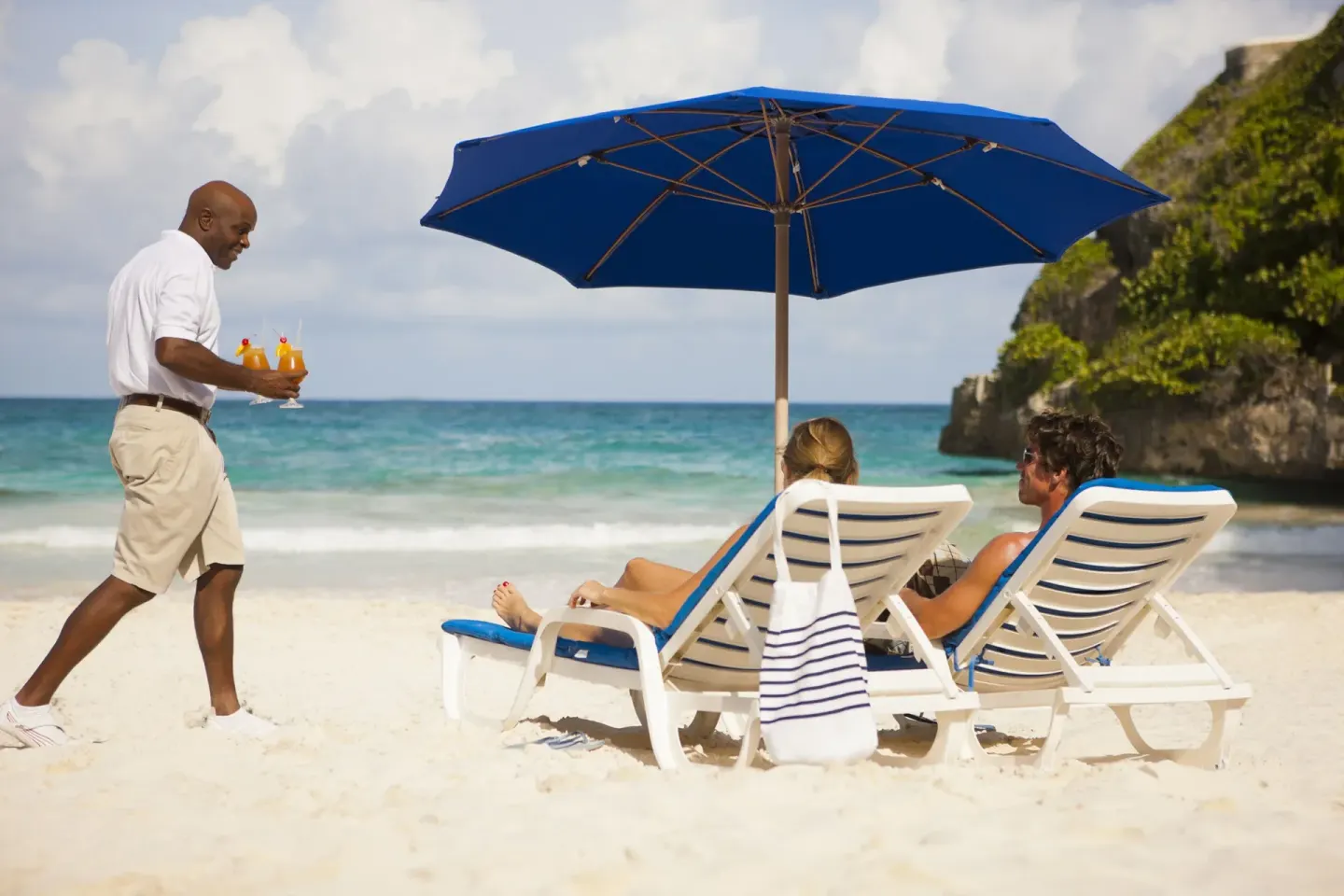 Beach loungers under a blue umbrella with drink service at The Crane Resort.