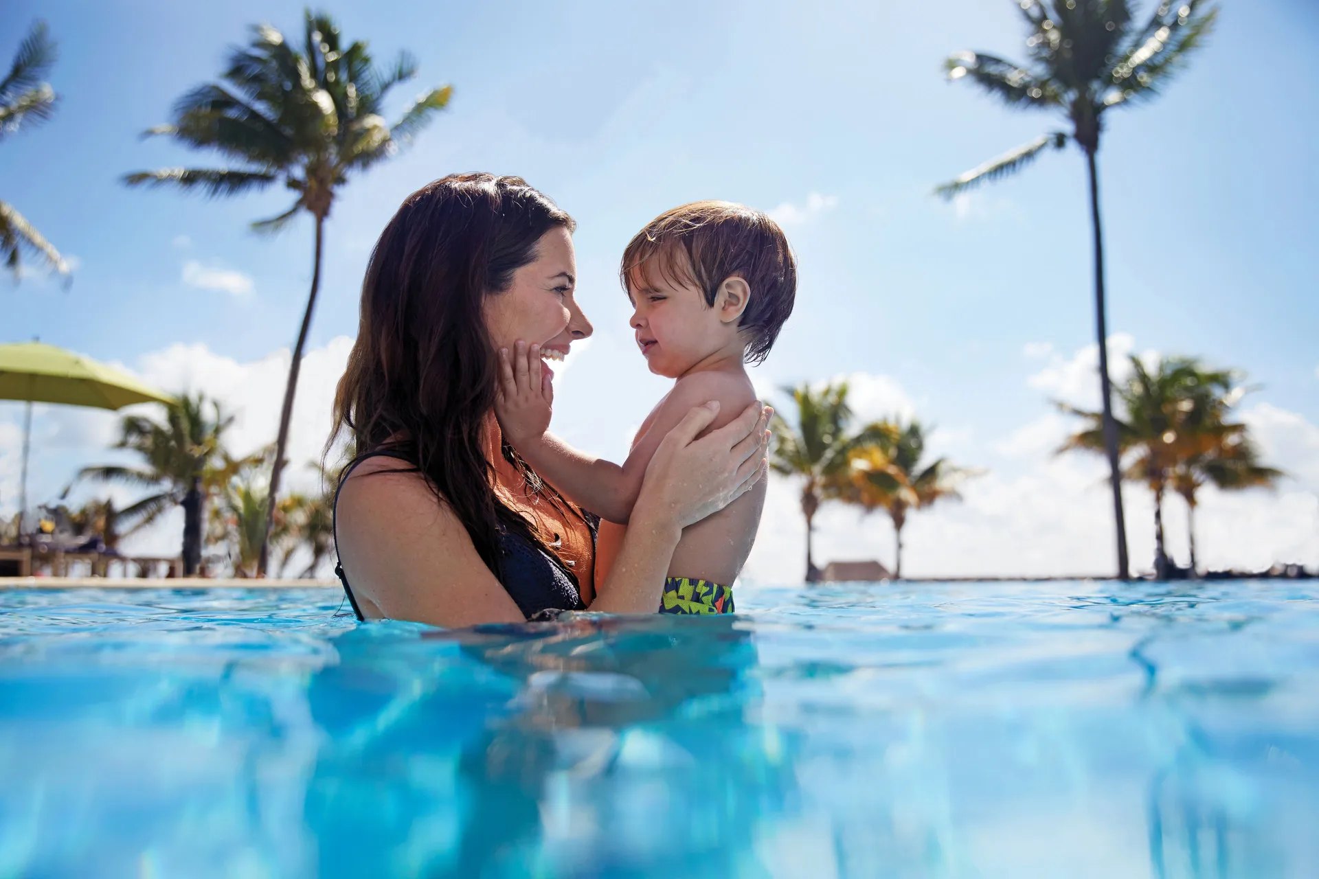 Adult holding a child in a palm-lined pool at The Fives Beach Hotel.