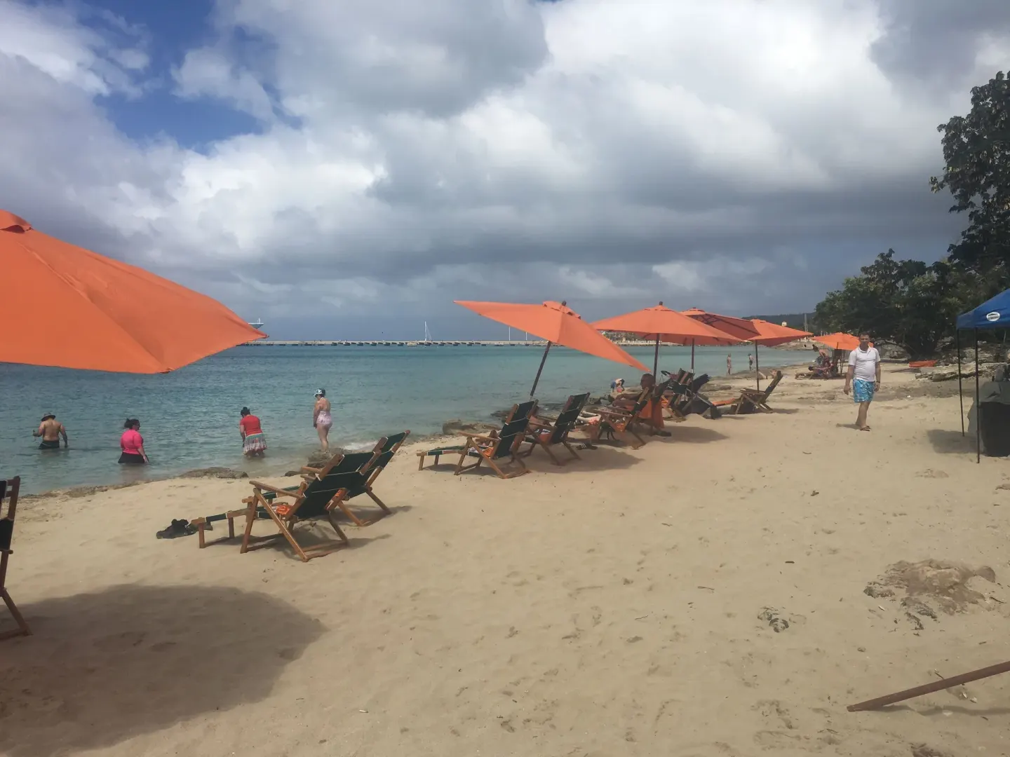 Sandy beach at The Fred with orange umbrellas, lounge chairs, swimmers, and cloudy sky.