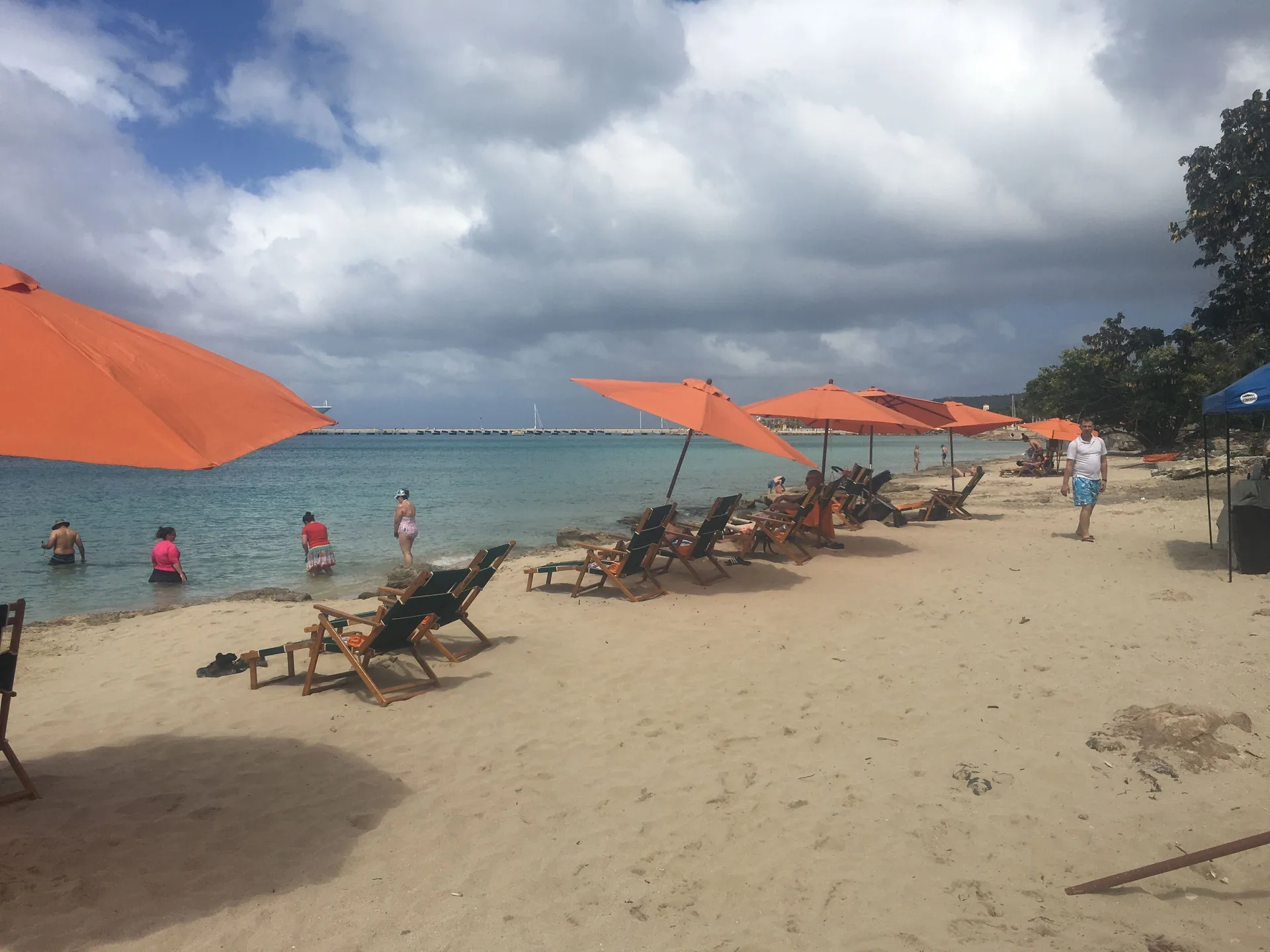 Sandy beach at The Fred with orange umbrellas, lounge chairs, swimmers, and cloudy sky.