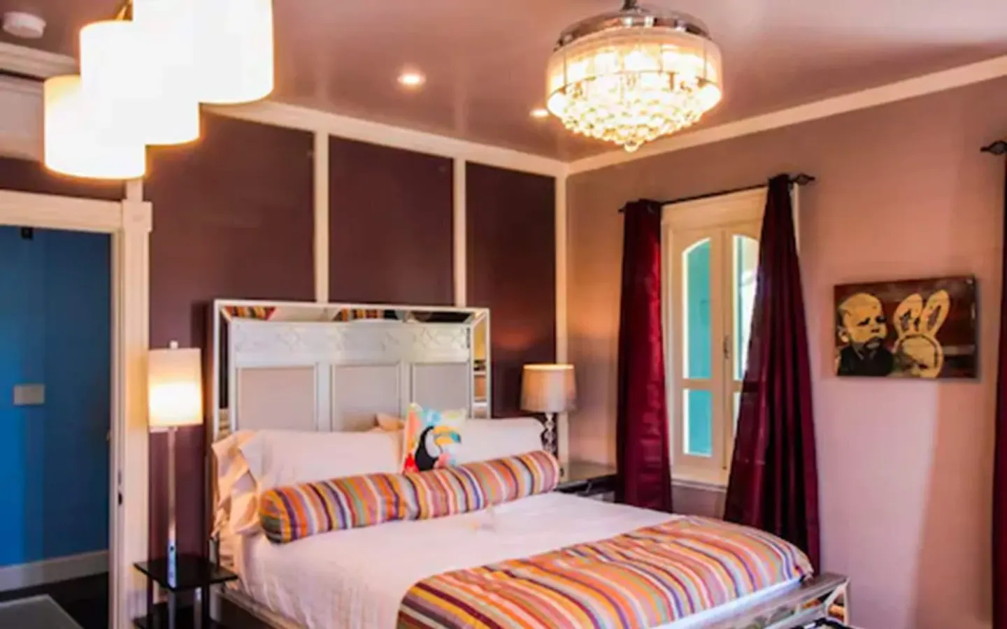 Colorful corner guest room with bed, chandelier, and tall windows at The Fred.