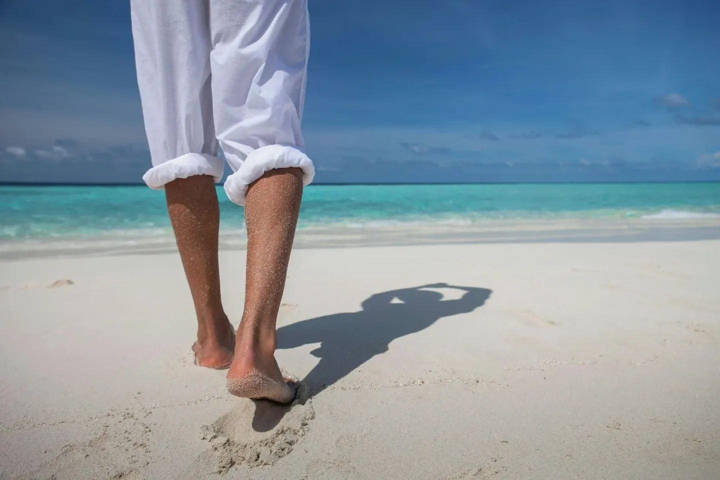 Barefoot person walking on white sand toward turquoise water at The House.