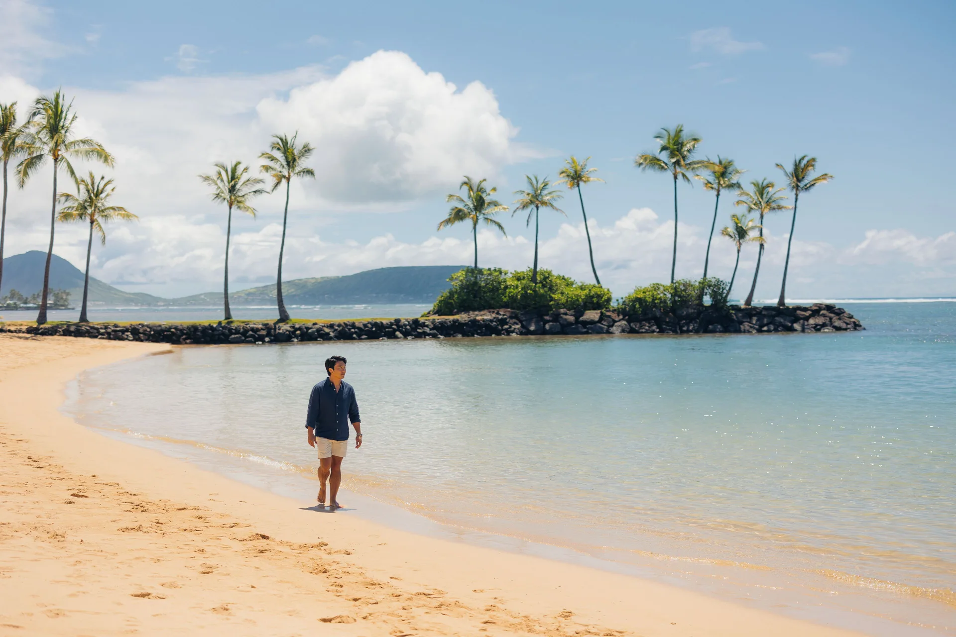 Man walking on the beach beside a palm-lined islet at The Kahala Hotel & Resort.
