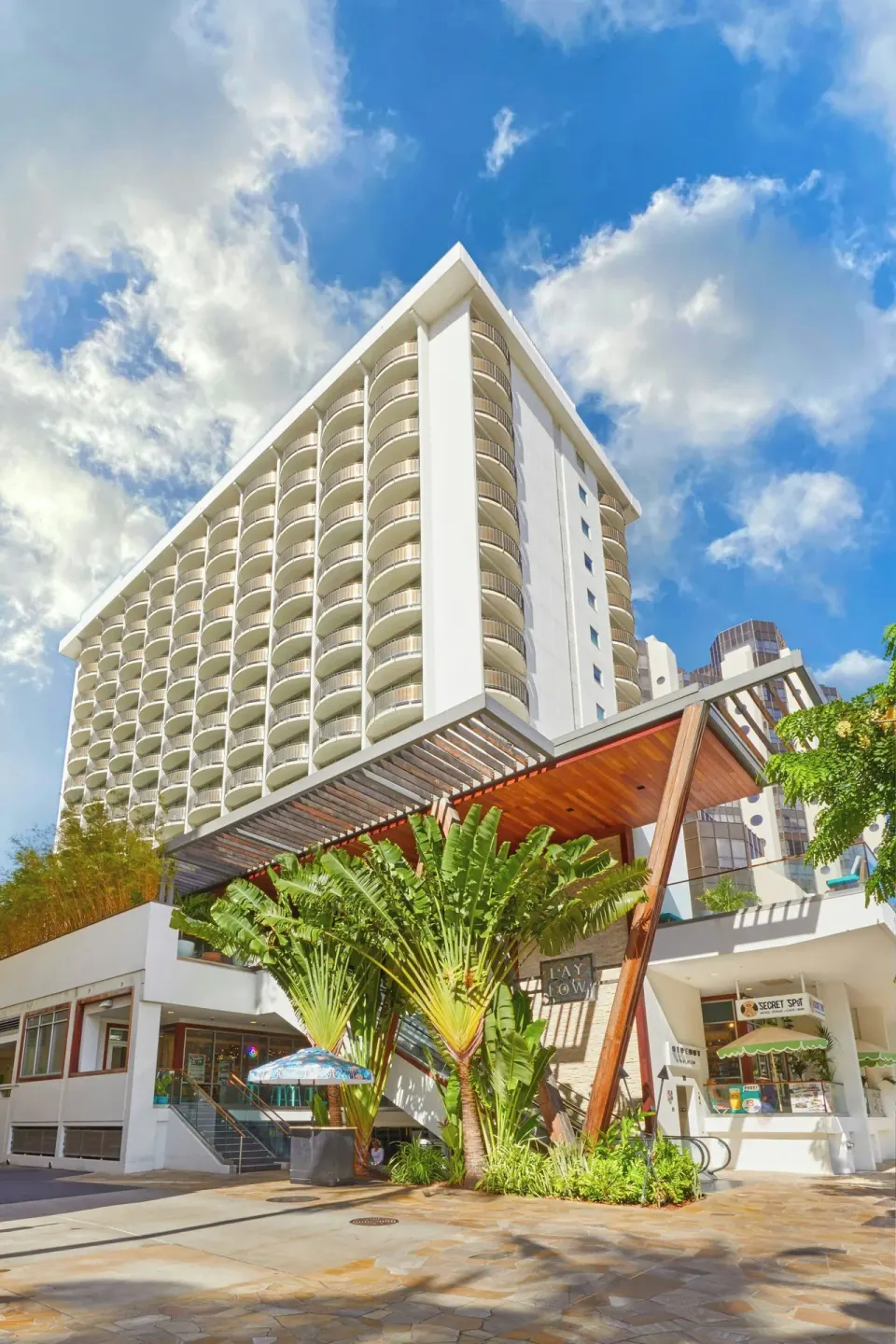 Street-level exterior of The Laylow Waikiki, Autograph Collection with palms and the hotel tower above.