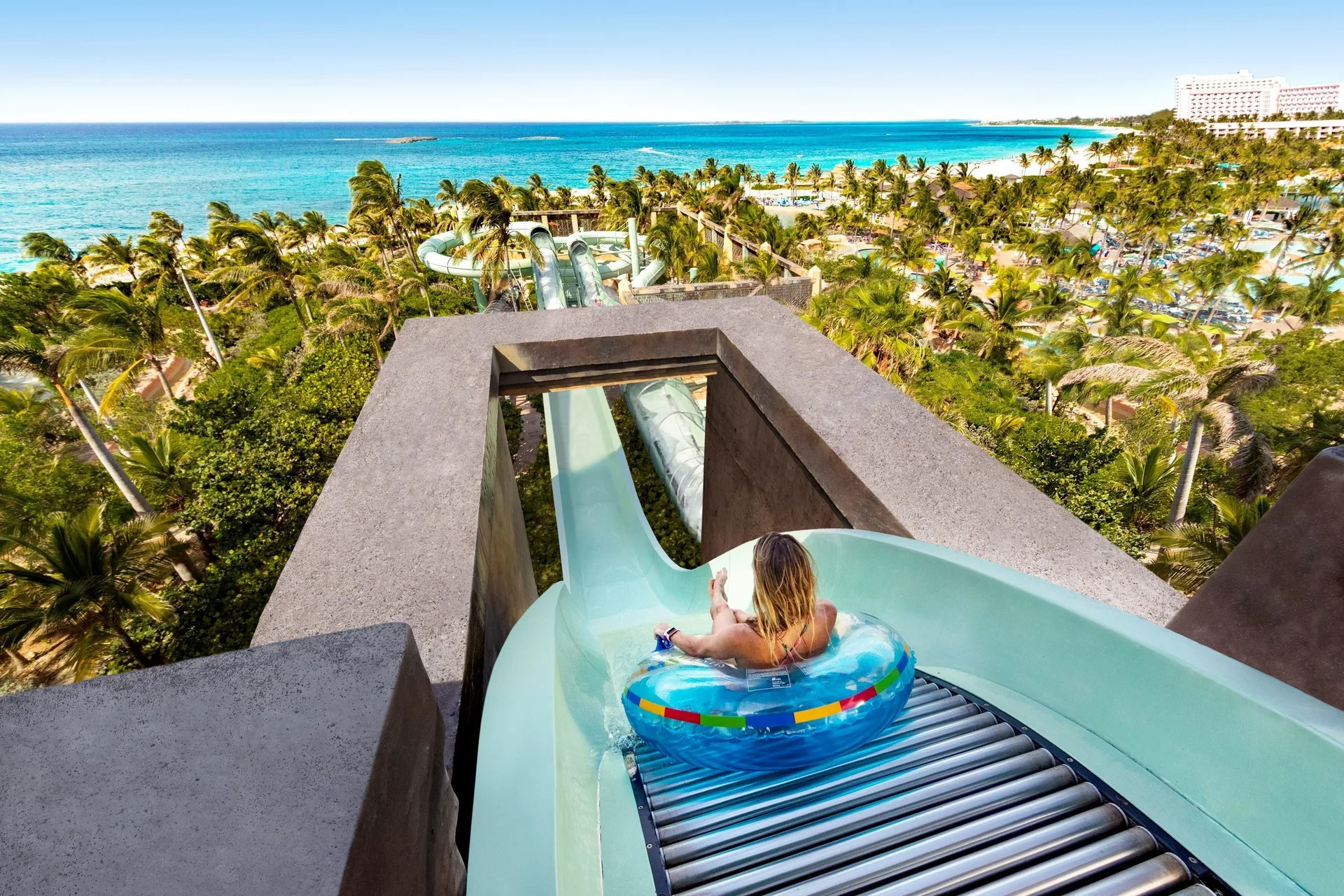 A rider on a tube enters a waterslide at The Reef at Atlantis with ocean and palms beyond.