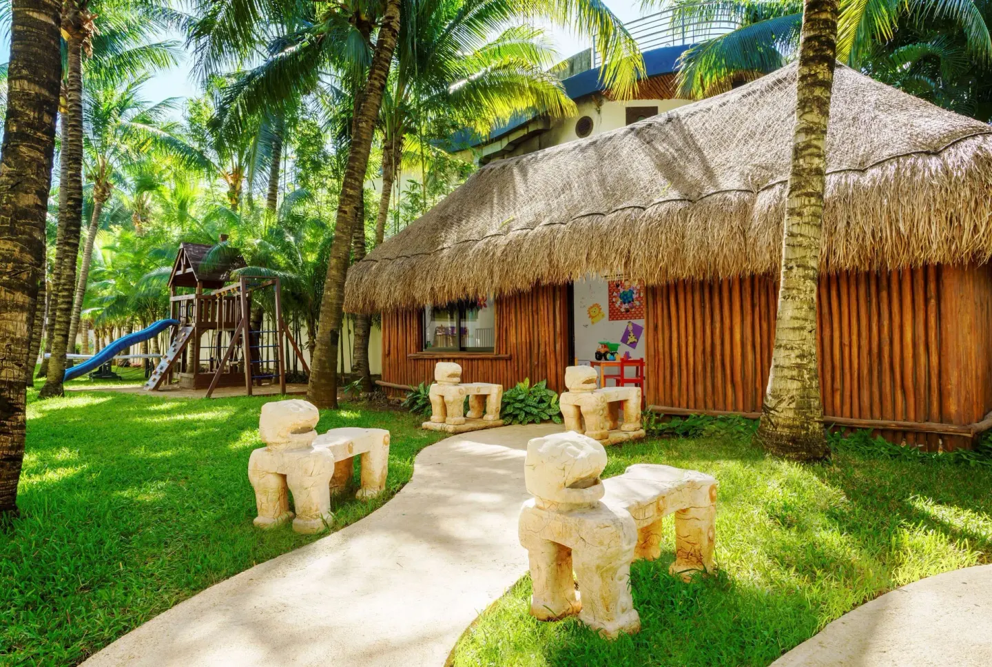 Palm-shaded kids' play area and thatched building at The Reef Coco Beach Resort & Spa.